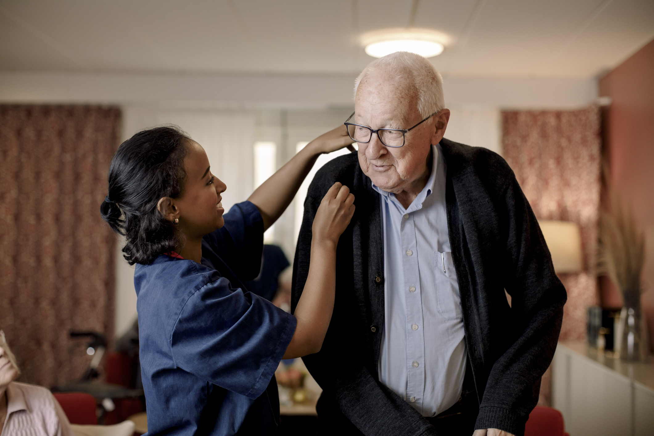 Senior man looking at female nurse helping him to wear sweater in retirement home