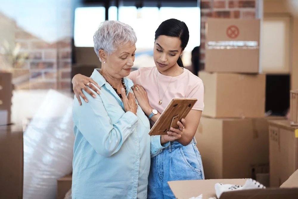 Older woman and young woman look at framed photo in room surrounded by boxes